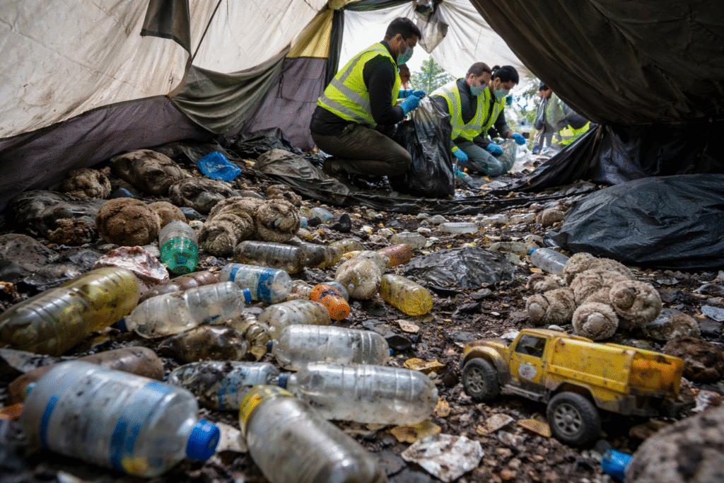 Cleanup workers in protective gear inside an abandoned Seattle encampment tent, surrounded by urine-filled bottles, drug debris, rotting trash, and children’s toys during a hazardous cleanup operation.