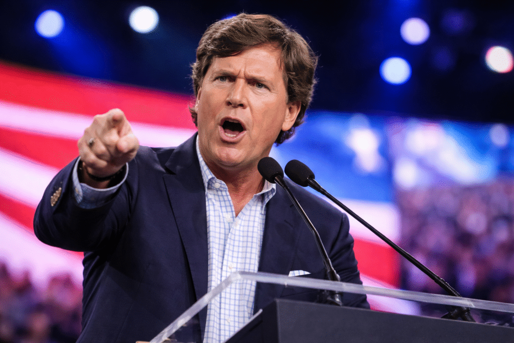 Tucker Carlson speaking at a political conference, pointing while standing at a lectern with microphones, wearing a navy blazer and light blue shirt, with an American flag backdrop behind him.