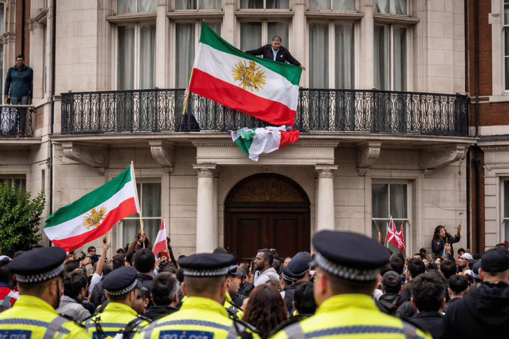 Protesters gather outside the Iranian embassy in London as an activist displays Iran’s pre-1979 royal flag from the balcony, with police officers and a crowd visible below.