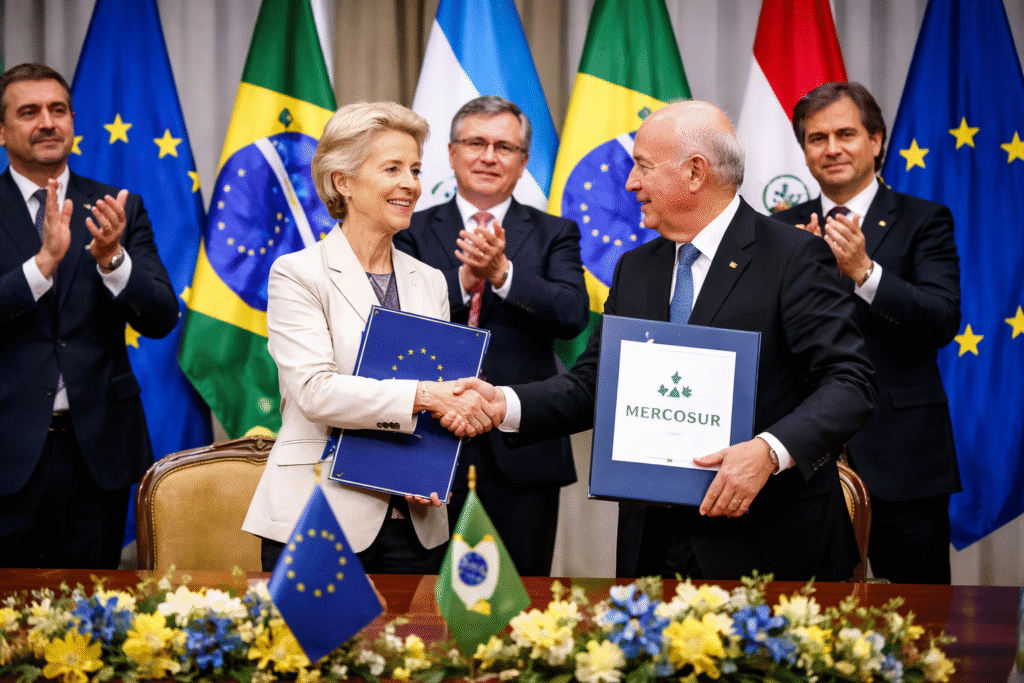 European Commission President Ursula von der Leyen and a Mercosur representative shake hands while holding signed trade agreement documents, with EU and South American flags displayed behind them.