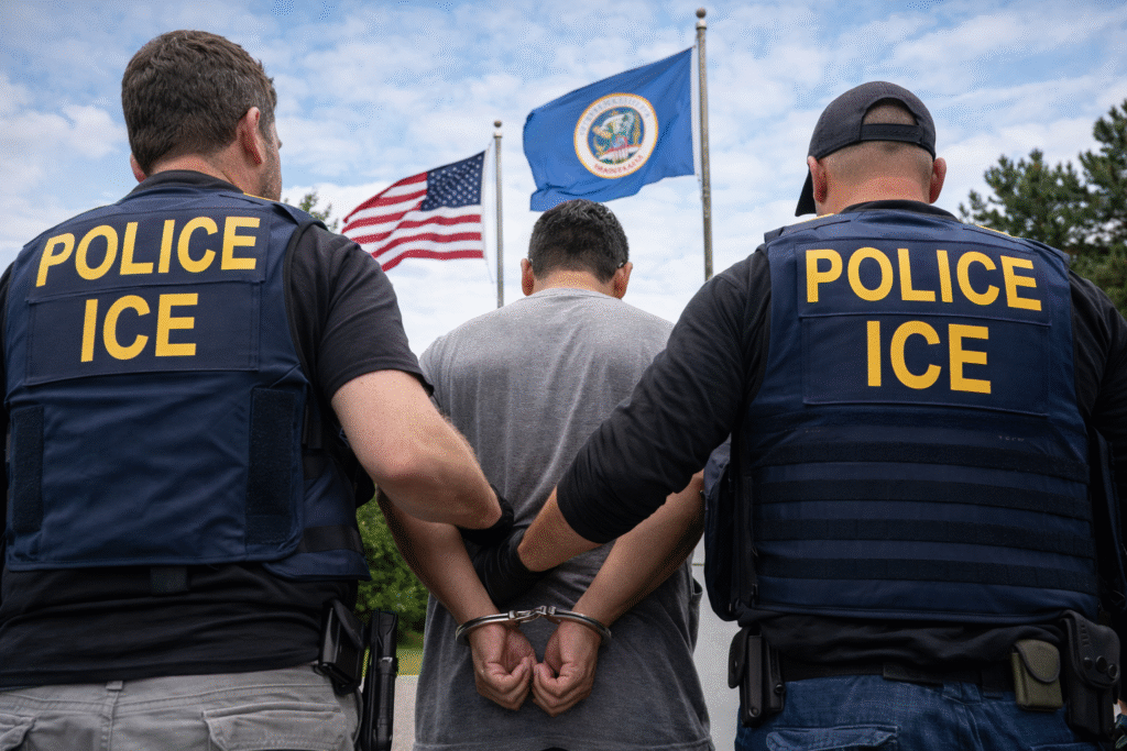 Two ICE agents in tactical vests escort a handcuffed man outdoors, with the Minnesota state flag and U.S. flag visible in the background.