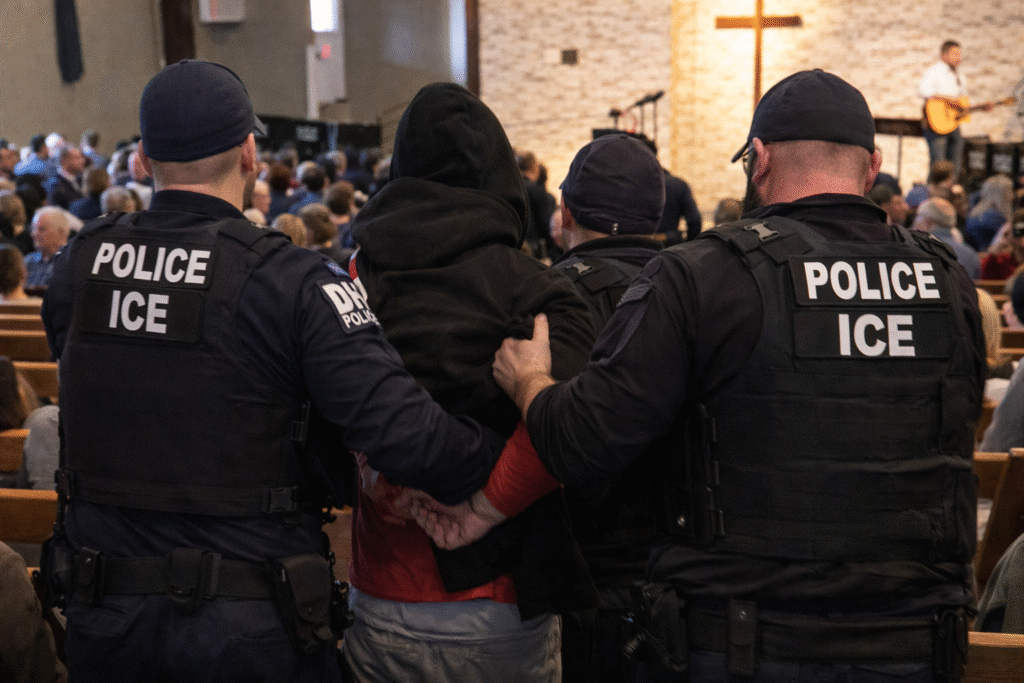 U.S. Immigration and Customs Enforcement officers escort a detained protester through a church sanctuary while congregants remain seated, with wooden pews, a cross, and a worship band visible in the background.