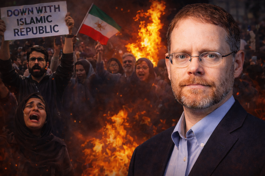 Daniel Greenfield stands in the foreground wearing a blazer and glasses, with Iranian protesters behind him holding flags and protest signs amid flames and smoke during a demonstration against Iran’s Islamic regime.