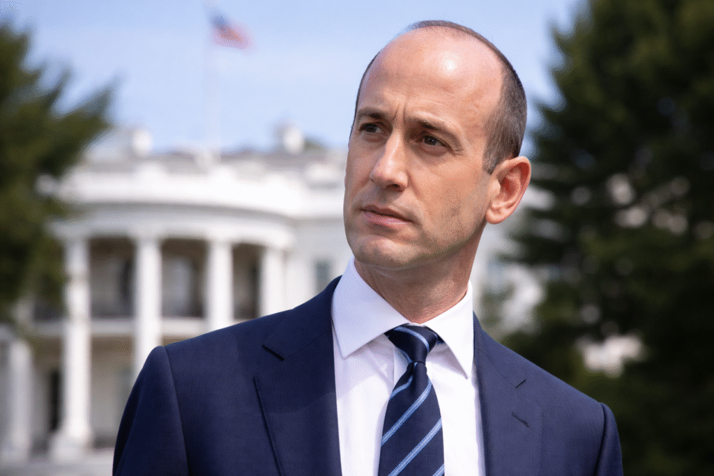Stephen Miller standing outside the White House, wearing a navy suit and blue striped tie, with the White House building blurred in the background on a clear day.