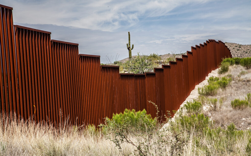 Steel border wall running through a desert landscape along the U.S.–Mexico border, with rust-colored fencing, sparse vegetation, and a cactus visible under a clear sky.