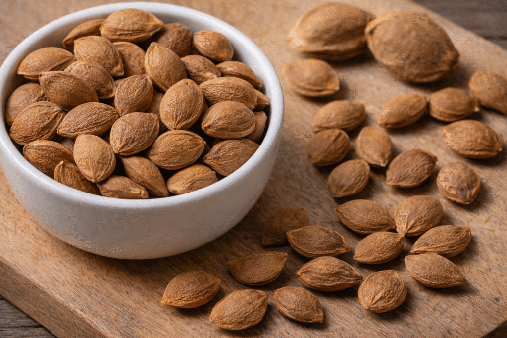 Close-up of raw apricot seeds (kernels) in a bowl and on a wooden board, showing the hard brown seed inside the fruit pit with a focus on their texture and size.
