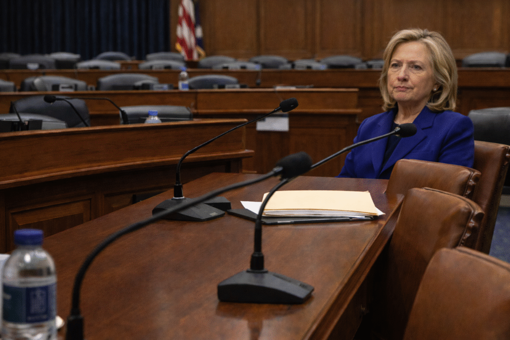Photograph of a U.S. congressional hearing room with microphones and empty chairs, representing controversy over a deposition photo from the Epstein inquiry involving Hillary Clinton.