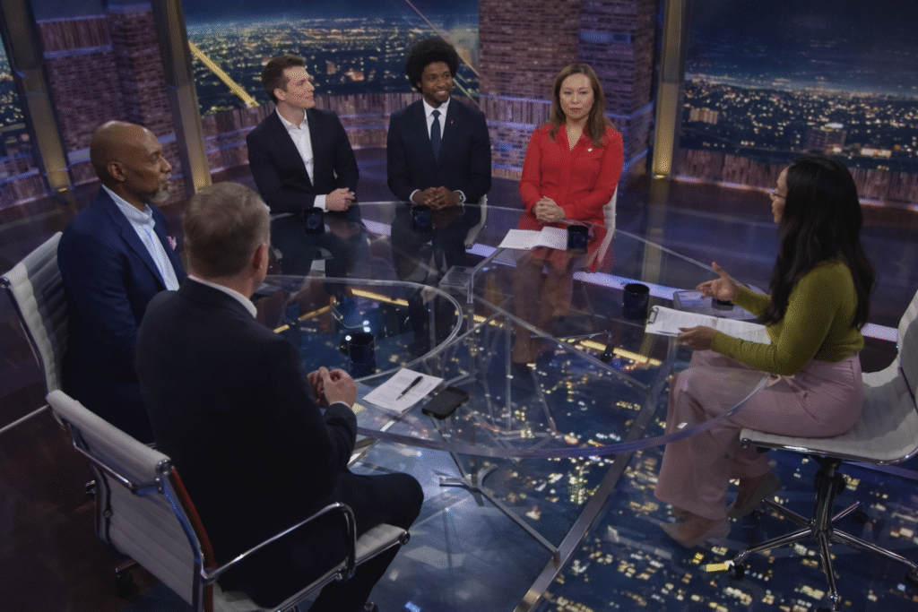 Six television panelists seated around a glass table in a studio with a nighttime city skyline backdrop, engaged in a live discussion.