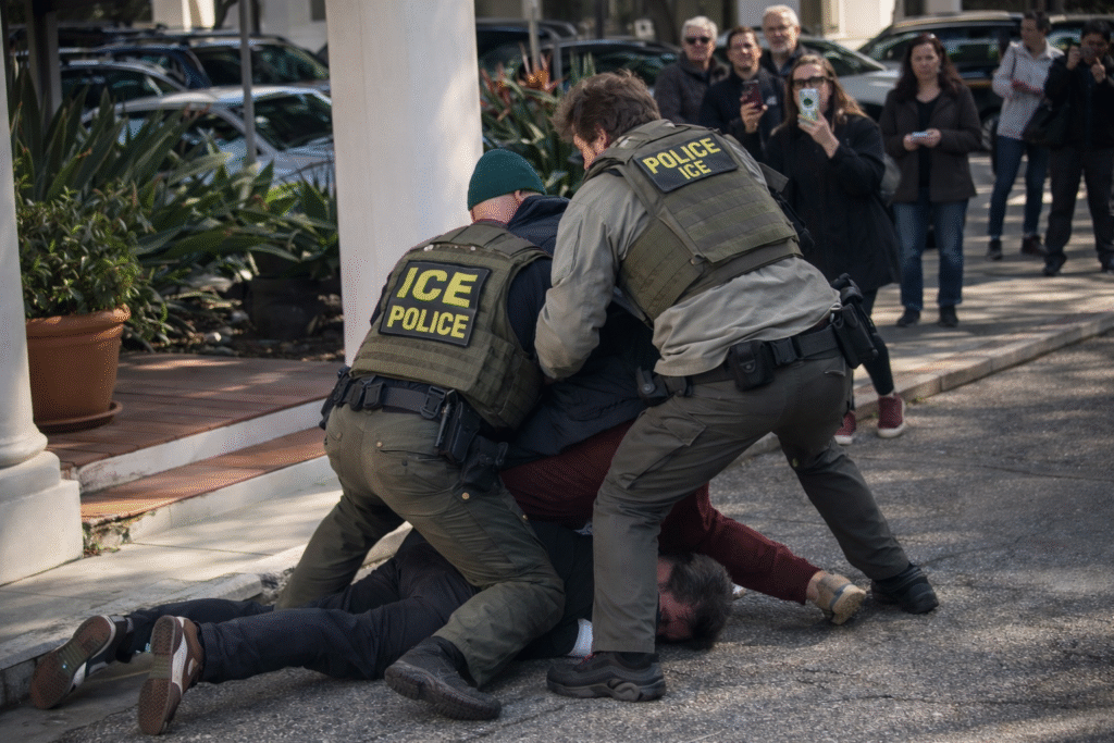 Two uniformed federal agents restraining a man on the ground outside a columned government building while several bystanders watch and record the incident.