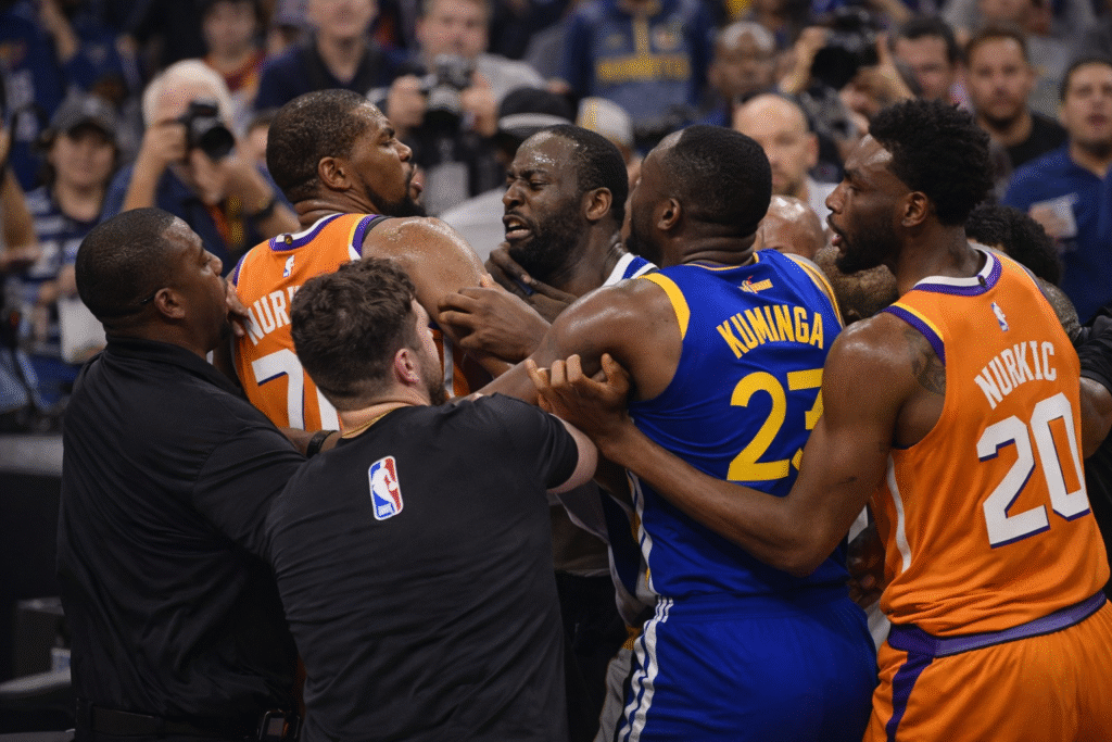 Players and staff from opposing teams restrain one another during a heated on-court altercation at a professional basketball game, with security intervening as the crowd watches in the background.