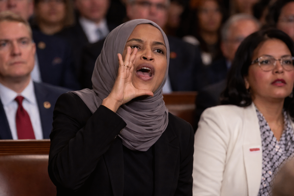 Rep. Ilhan Omar wearing a gray hijab raises her hand and shouts while seated in the House chamber during a State of the Union address, with other lawmakers visible behind her.
