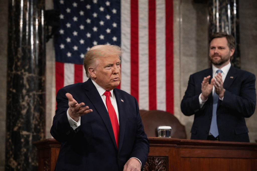 President Donald Trump gestures with an open hand while speaking in the U.S. Capitol House chamber, with Senator JD Vance standing behind him applauding and an American flag displayed in the background.