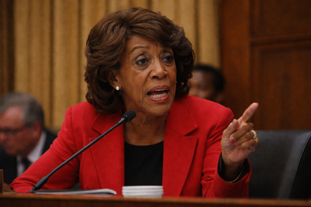 Representative Maxine Waters speaking into a microphone during a House hearing, wearing a red blazer and gesturing as she addresses the committee.