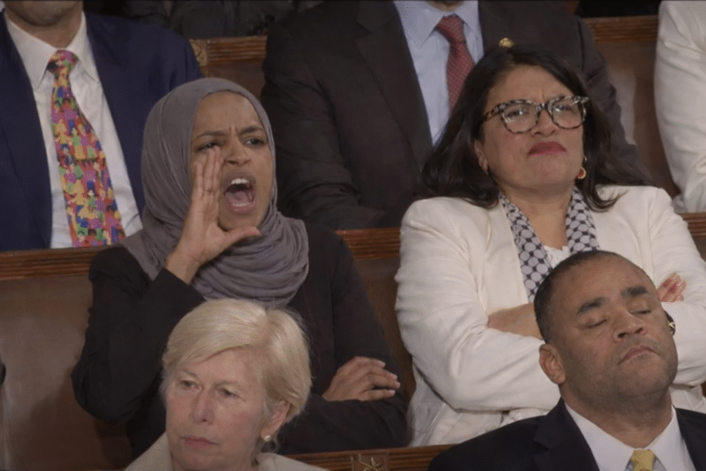 Close-up of a congresswoman in a gray hijab seated in a legislative chamber, cupping her hand to her mouth as she shouts, while another congresswoman beside her sits with arms crossed and a stern expression.