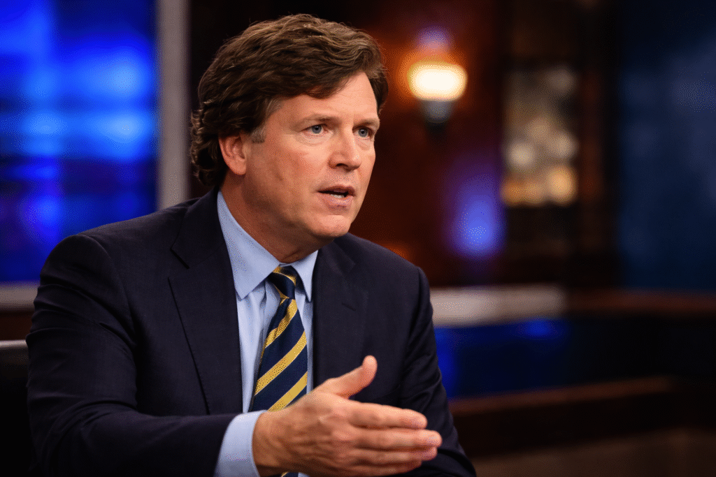 Tucker Carlson seated in a television studio, wearing a dark suit and striped tie, speaking while gesturing with his hand.