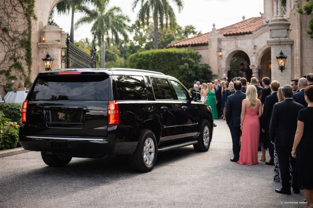 Image of a black SUV arriving at a Palm Beach estate entrance with well-dressed guests standing nearby in neutral daylight at a wedding event.