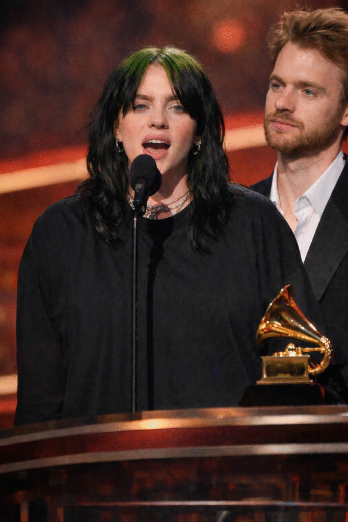 Billie Eilish speaks into a microphone at the Grammy Awards podium with her brother Finneas standing beside her, as a Grammy trophy rests on the stage under warm lighting.