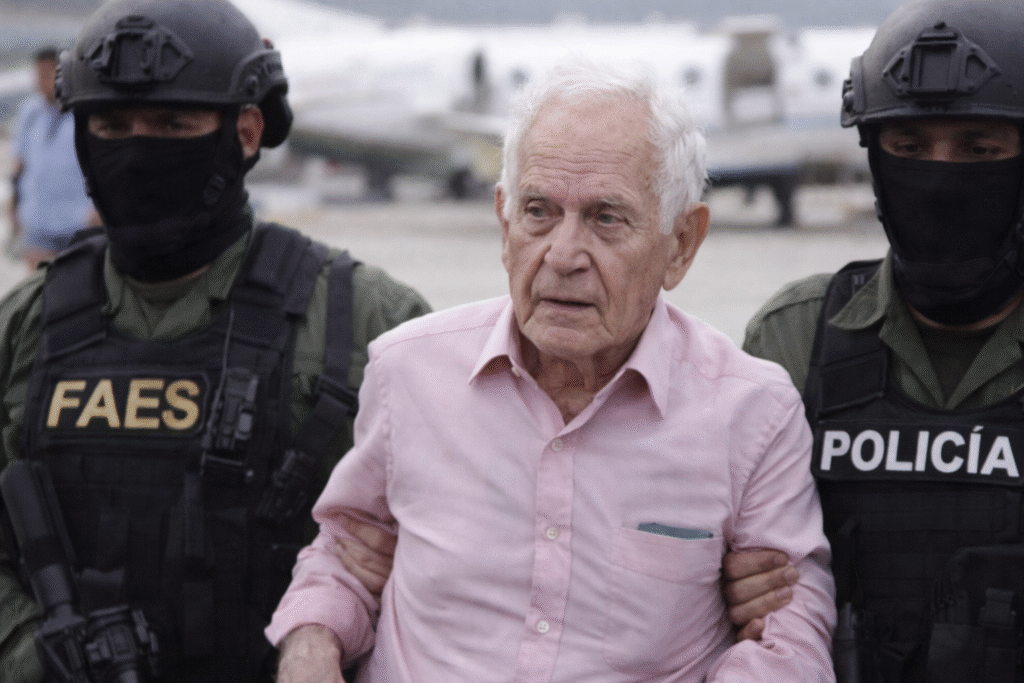 Elderly man escorted by armed Venezuelan officers on an airport tarmac with an aircraft in the background.