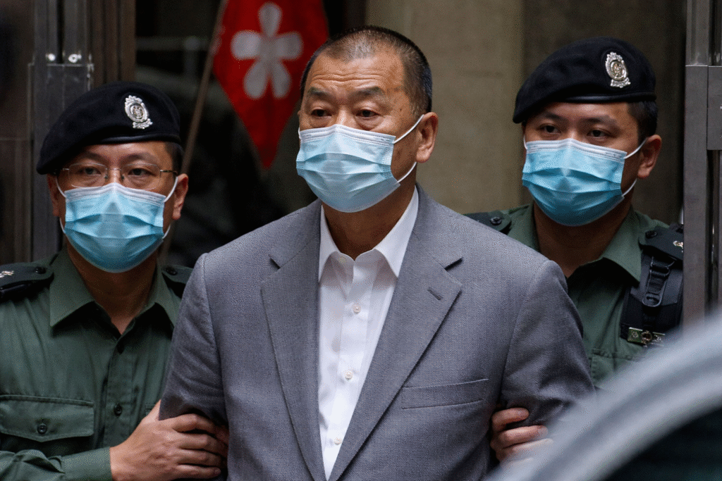 Jimmy Lai in a gray suit and face mask escorted by uniformed Hong Kong officers inside a government building.
