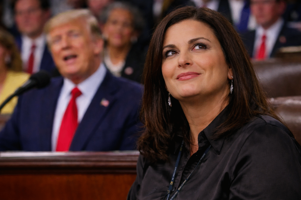 Woman with shoulder-length dark brown hair seated in the audience at a State of the Union address, glancing upward with a subtle expression as President Donald Trump speaks in the blurred background.
