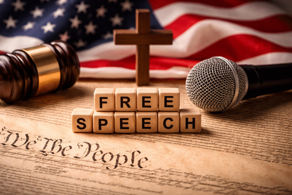 Close-up of the U.S. Constitution with a gavel, microphone, wooden cross, and American flag in the background symbolizing debate over free speech, religion, and law.