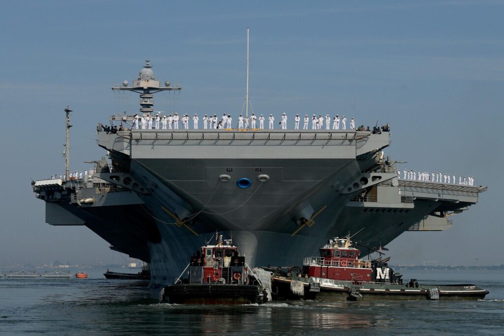 USS Gerald R. Ford aircraft carrier sailing with escort ships in open waters as fighter jets launch from the flight deck during naval operations.