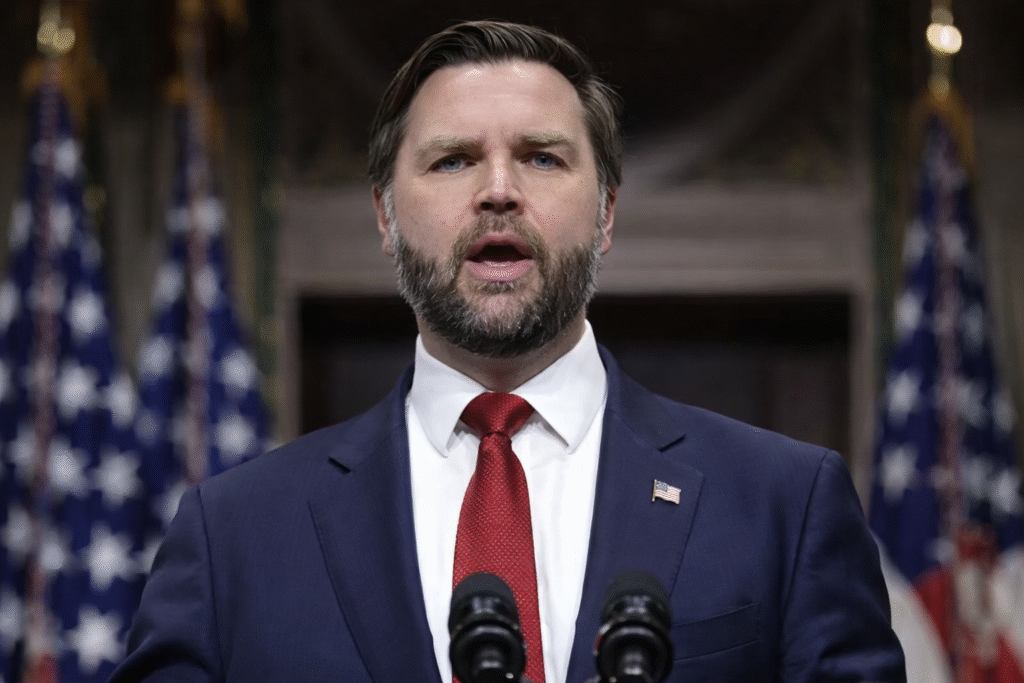 J.D. Vance standing at a podium with microphones, wearing a navy suit and red tie, speaking in front of American flags in a formal government setting.