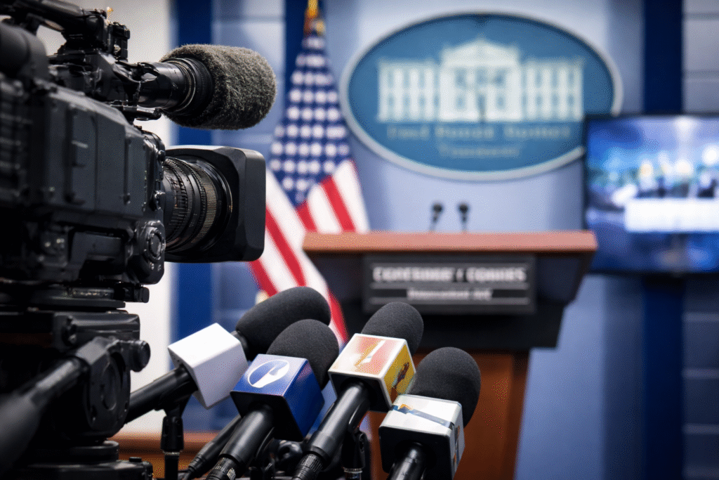 Television camera and news microphones focused on the White House press briefing room lectern with a blurred background screen symbolizing media attention and political commentary.