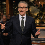 Late-night talk show host speaking on a studio stage during a political discussion in front of a live audience.