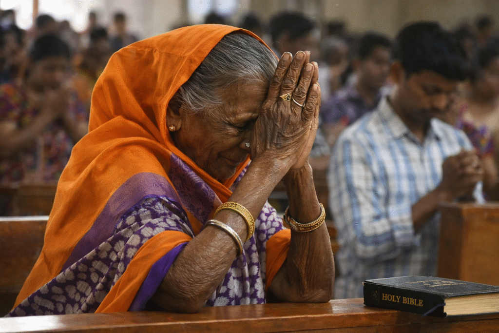 Elderly South Asian woman in an orange headscarf and patterned sari praying with hands clasped inside a church, with a Bible resting on the pew beside her and other worshippers blurred in the background.