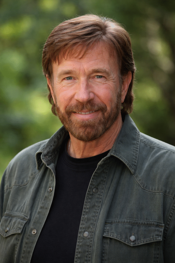 Chuck Norris smiling outdoors in a close-up portrait, wearing a dark green jacket over a black shirt with a blurred natural background