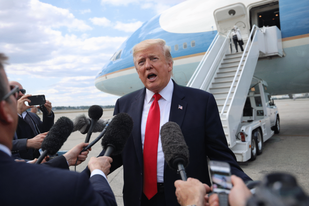 Donald Trump speaking to reporters on an airport tarmac with microphones extended toward him, standing in front of a large aircraft with boarding stairs visible behind him