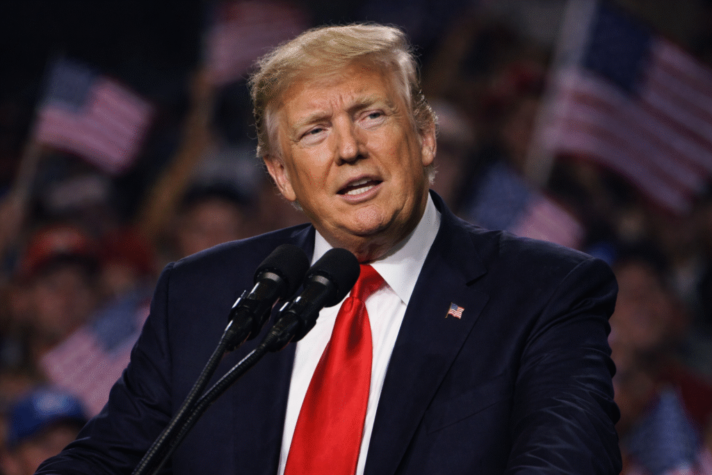 Donald Trump speaking at a podium with microphones, wearing a dark suit and red tie, with a blurred crowd in the background.