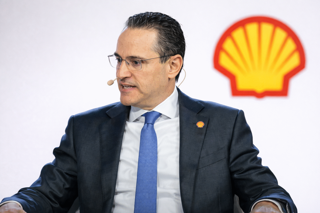 Middle-aged man in a dark suit and blue tie speaking at a conference, seated against a bright backdrop with an energy company logo behind him.