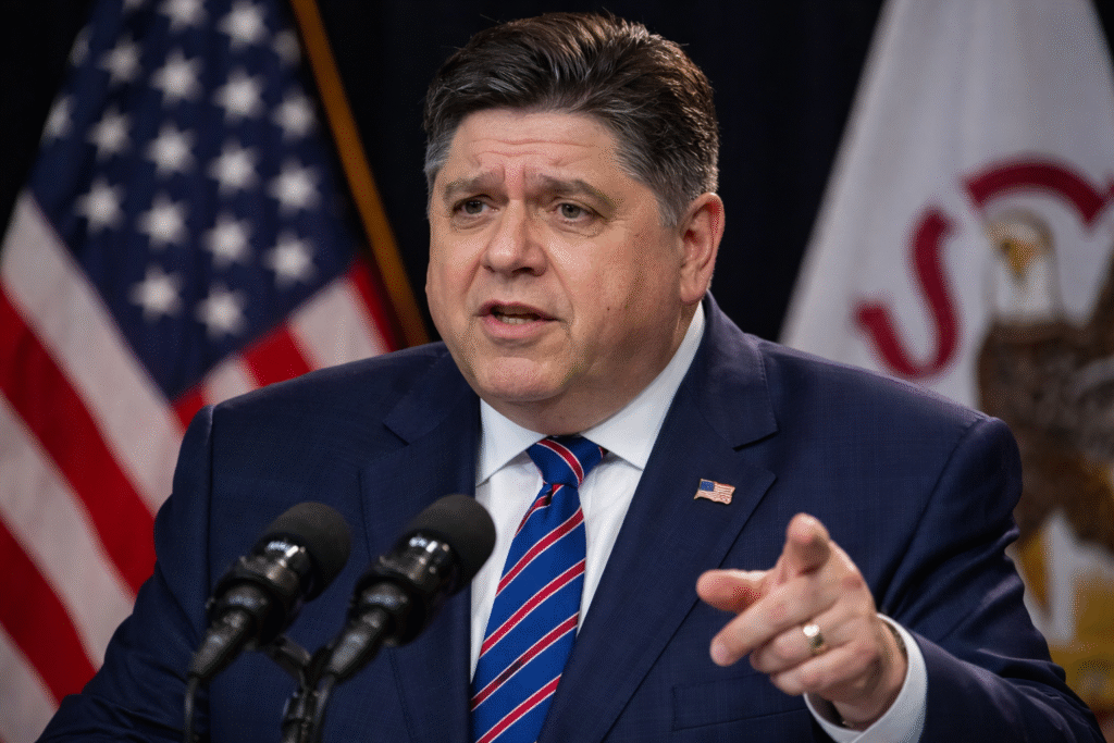 Illinois governor speaking at a press conference with microphones, gesturing while standing in front of U.S. and state flags