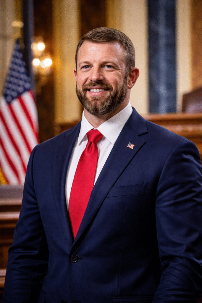 Markwayne Mullin standing in a formal government setting wearing a navy suit and red tie, with an American flag visible in the background