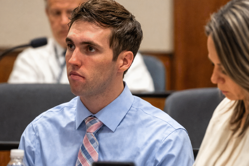 Young man in a blue dress shirt and striped tie seated in a courtroom during a legal hearing, with an attorney beside him reviewing documents.