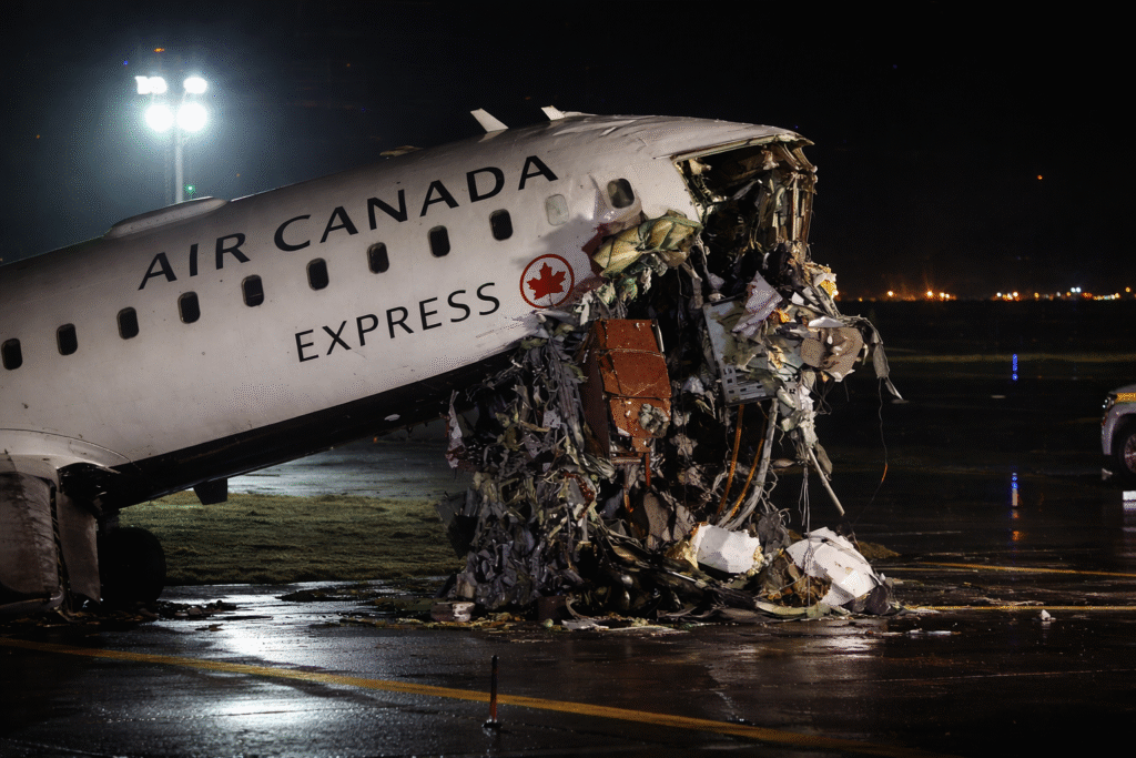 A heavily damaged passenger aircraft nose rests on a wet runway at night, with debris and exposed internal components visible under bright emergency lights.