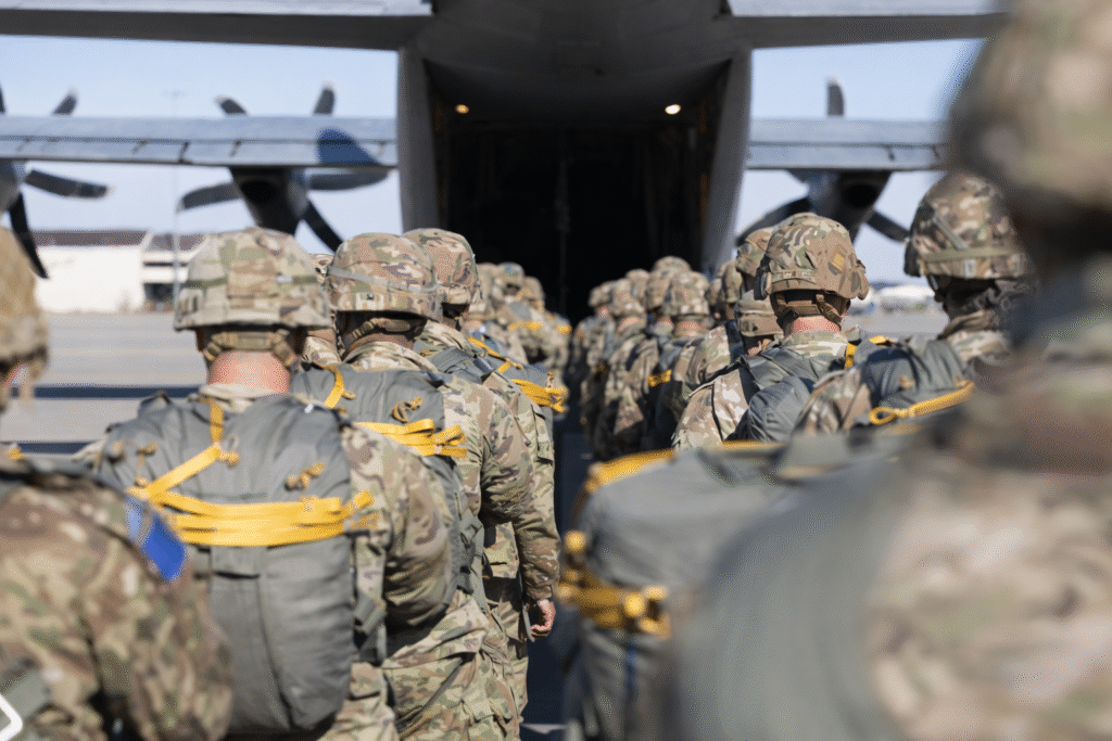 Soldiers in camouflage uniforms and parachute gear walking in formation toward a military transport aircraft with its rear ramp open on an airfield.
