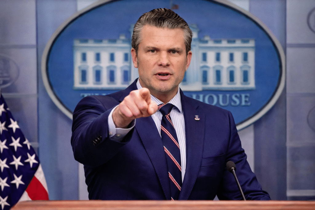 Pete Hegseth speaking at a White House press briefing podium, pointing forward with a serious expression, with the White House emblem visible in the background.