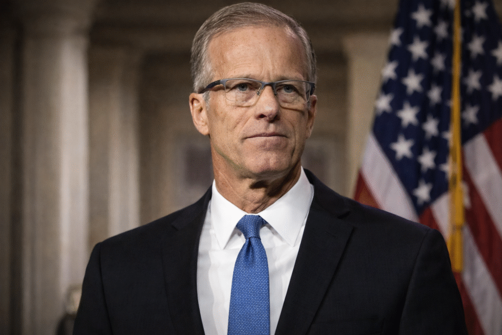 A middle-aged man in a dark suit and blue tie stands at a podium with a serious expression, speaking in a formal setting with an American flag in the background.