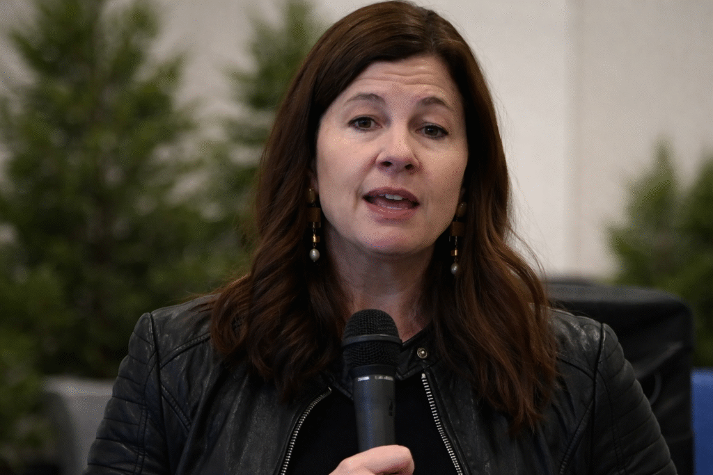 Woman speaking into a handheld microphone indoors with a blurred neutral background and no visible text or signage.