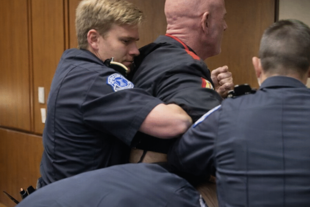 Capitol Police officers restrain and escort a bald protester from a wood-paneled hearing room during a congressional session while others assist nearby.