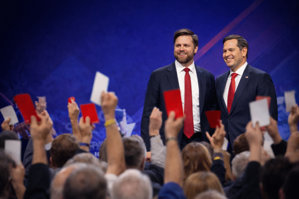 JD Vance and Marco Rubio standing together on stage in suits and red ties, smiling toward an audience holding up voting cards in a political event setting.