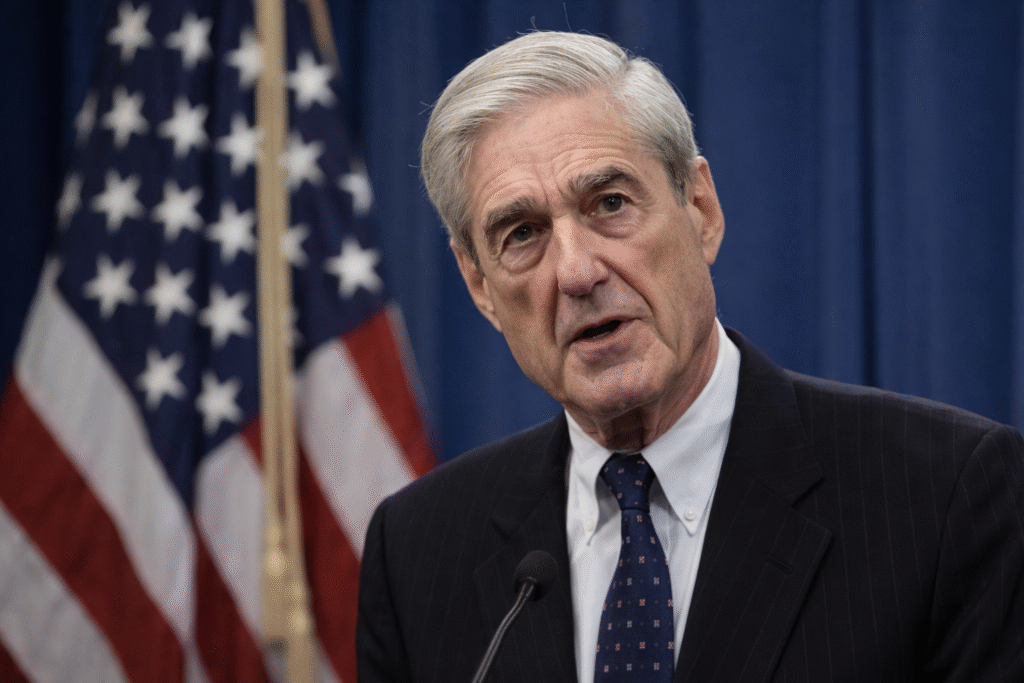 Robert Mueller speaking at a podium with an American flag behind him, wearing a dark suit and tie against a blue curtain backdrop.