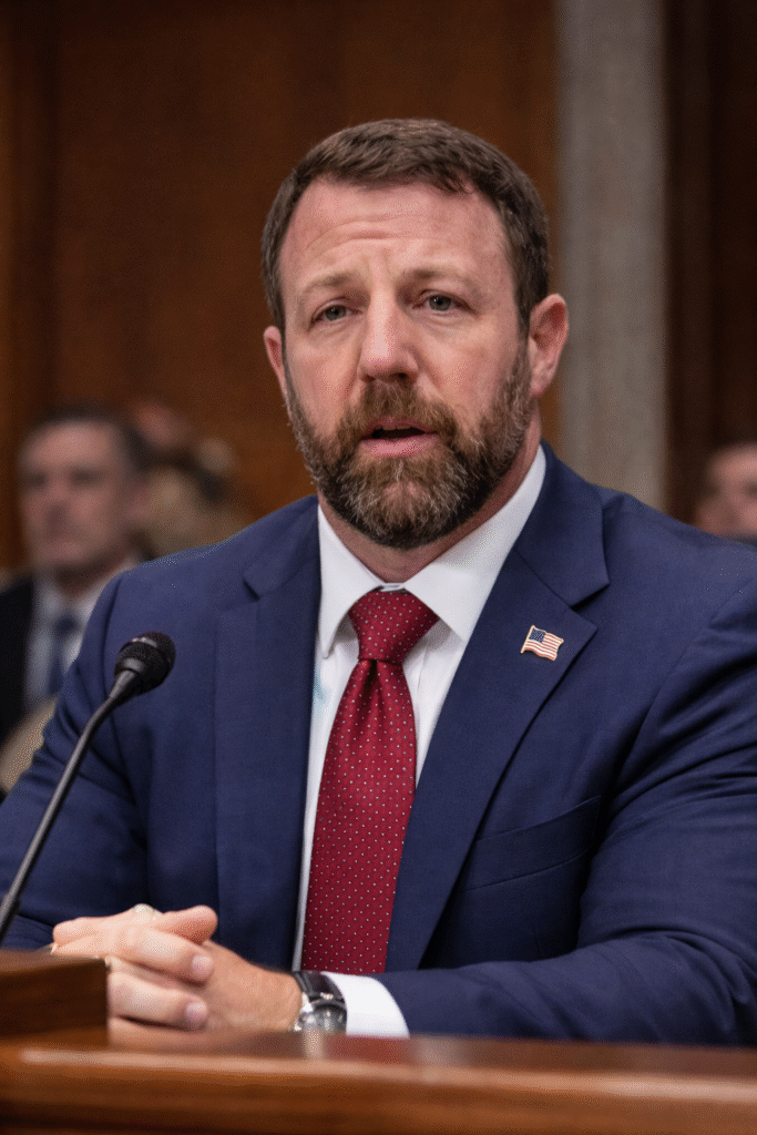 Markwayne Mullin speaking outdoors near the U.S. Capitol building, wearing a navy suit and red tie, looking to the side with government architecture in the background