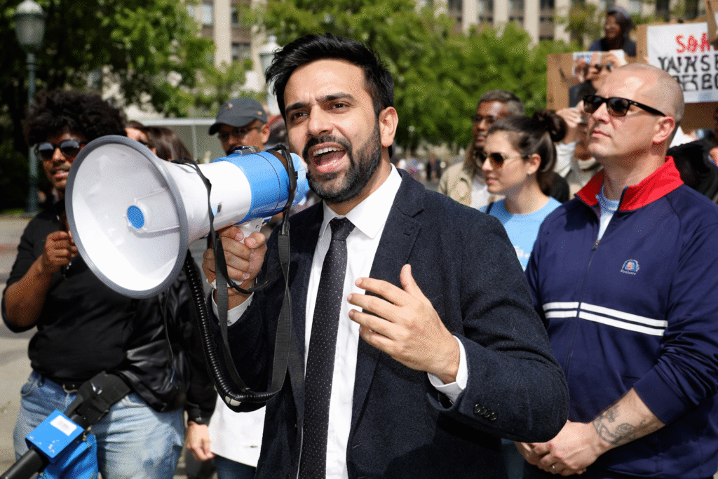 Protest speaker using a megaphone while addressing a crowd during a public demonstration in a city park with supporters and protest signs in the background.