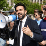 Protest speaker using a megaphone while addressing a crowd during a public demonstration in a city park with supporters and protest signs in the background.