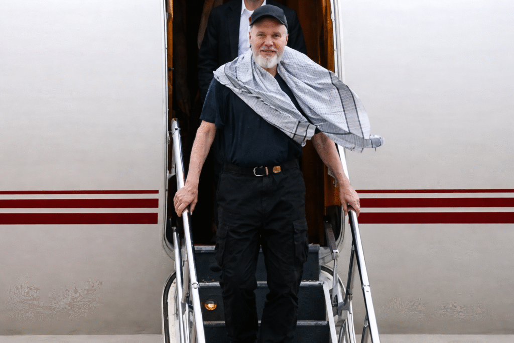 A man with a white beard wearing a dark cap and casual clothing descends the stairs of a private aircraft, with a scarf draped over his shoulders, in a calm airport setting.