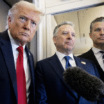 Former President Donald Trump speaking with reporters inside an aircraft doorway while two officials stand beside him as a microphone is held toward them.